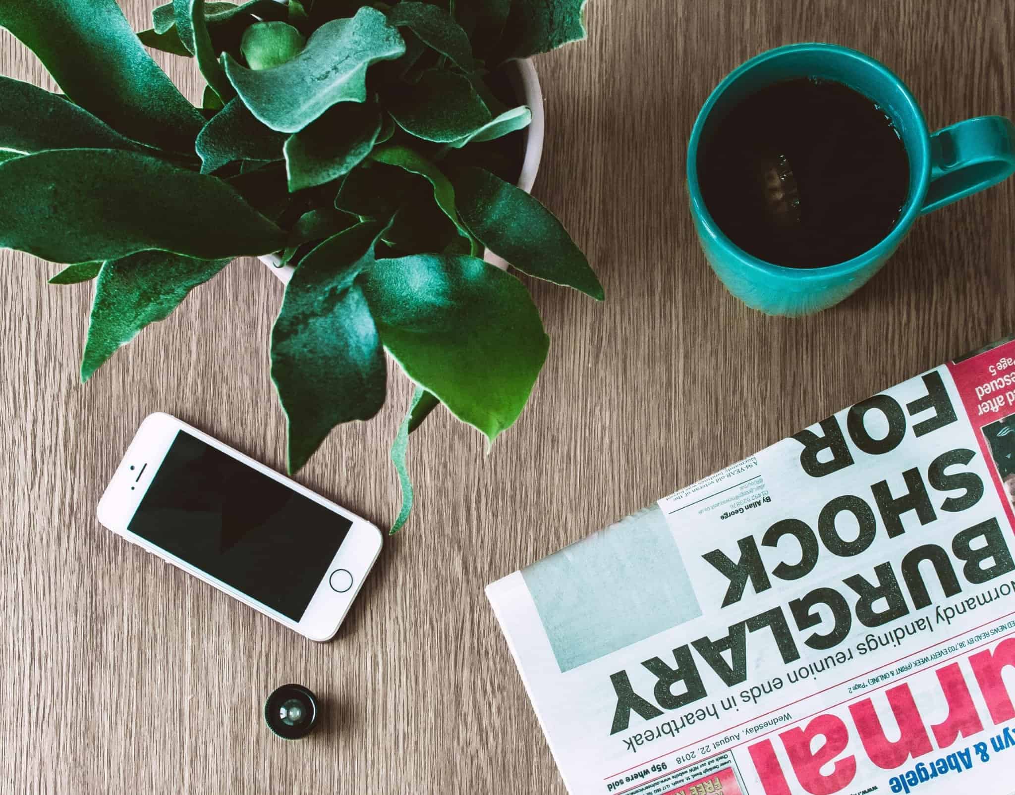 Top view of a wooden table with a green plant, a white smartphone, a teal coffee mug, and a folded newspaper